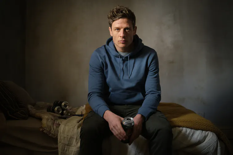 Tommy Lee Royce sits on a bed on a blue hoody, holding a can of beer. Five more are on the bed behind him.