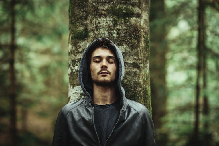 Young man in a forest leaning on a tree with eyes closed
