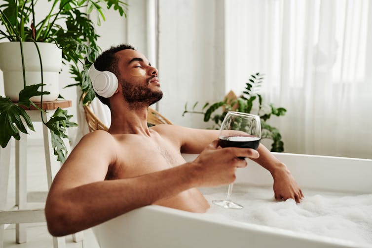 Man relaxing in bath with glass of wine surrounded by houseplants