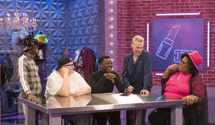 Five drag race contestants sit around a table in conversation. A neon poster of a lipstick hangs in the background.