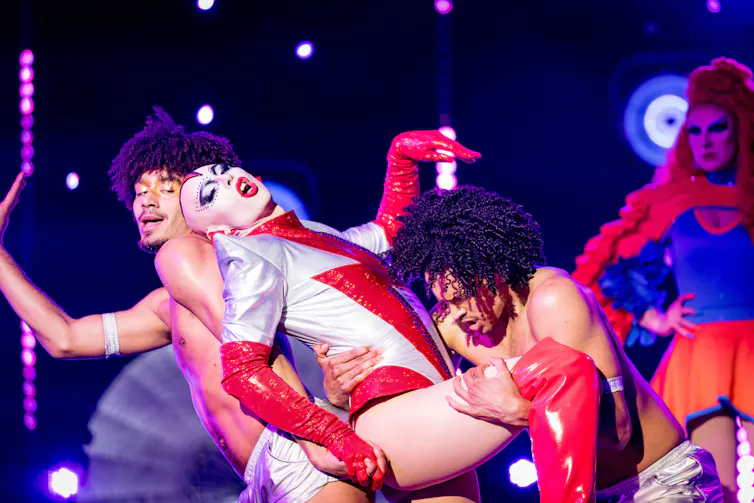 Cheddar Gorgeous dances in a silver and red leotard, with a red lightning bolt on the front. She is bald, with dramatic makeup and leaning on topless male dancers.