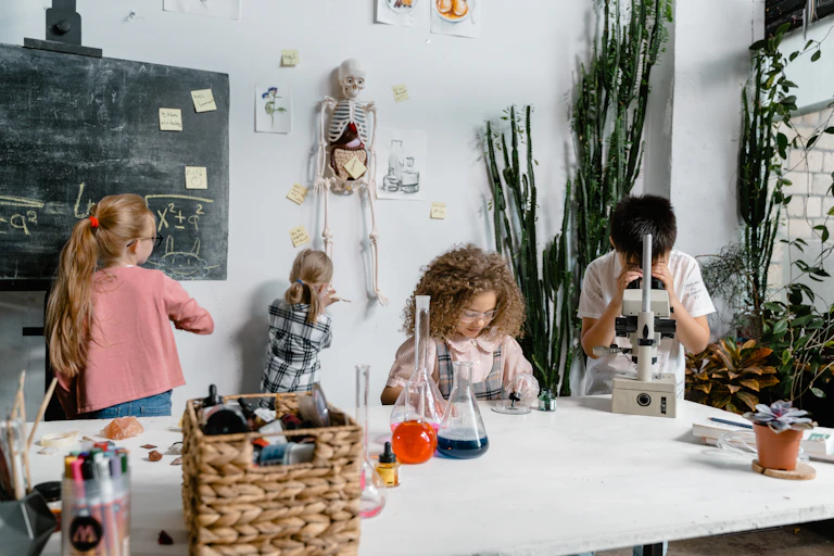 Children in a lab write on a blackboard and peer down a microscope