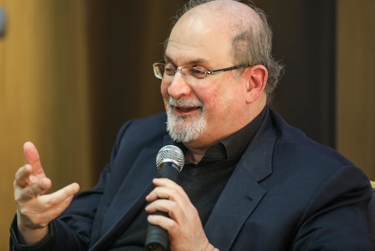 Salman Rushdie in a blue suit holding a microphone.