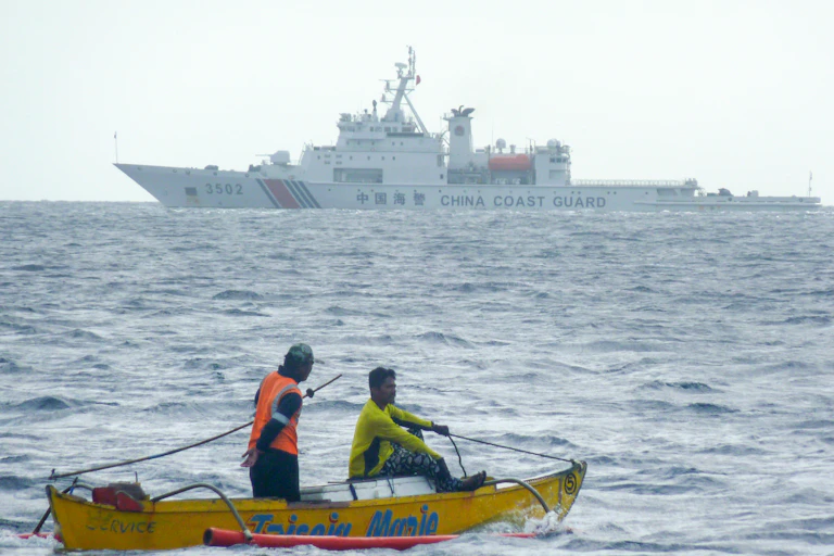 Deux pêcheurs sur une barque en mer. Au large, un navire militaire.