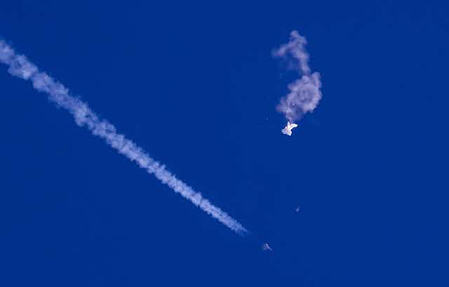 The remnants of a large white balloon drift from the sky, with a fighter jet and its contrail below it