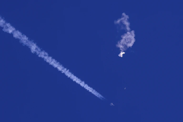 The remnants of a large white balloon drift from the sky, with a fighter jet and its contrail below it
