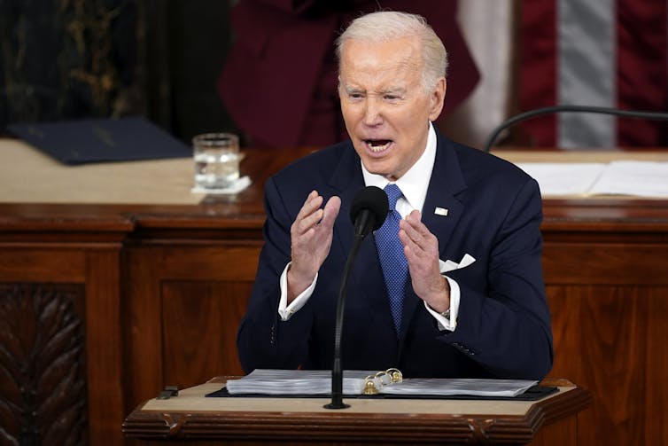 elderly white man with gray hair stands in front of lectern and appears to speak while gesticulating with his hands