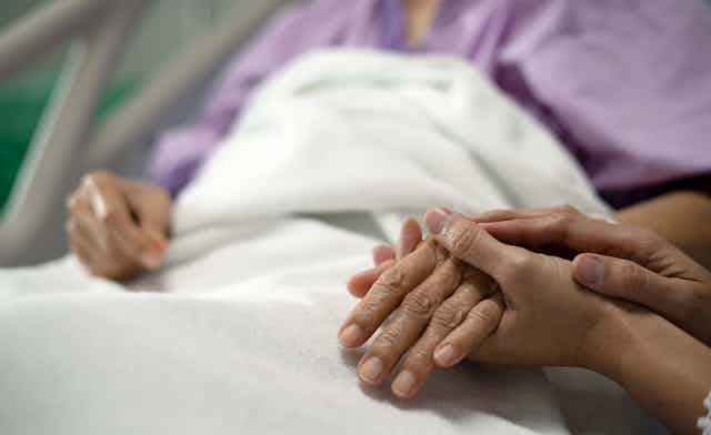 An older person of color in a hospital bed clasps hands with a younger person.
