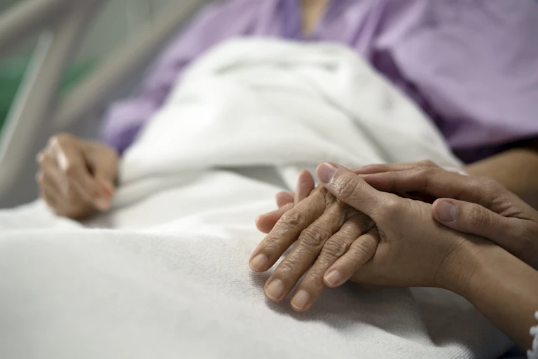An older person of color in a hospital bed clasps hands with a younger person.