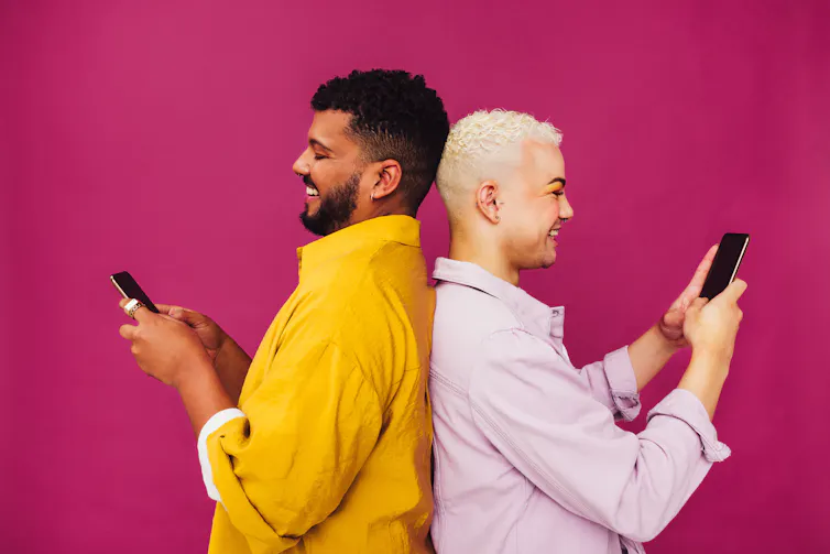 An androgynous couple, both with short hair and wearing makeup and brightly-coloured collared shirts, stand back to back and smile at their mobile phones, against a bright pink background