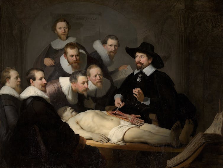 Six balding doctors gather round to watch a demonstration of a dissection.