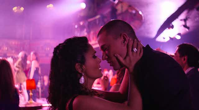 Channing Tatum and Salma Hayek Pinault almost kiss against a pink lit room.