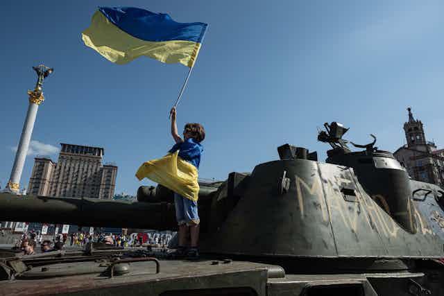 A small child is wrapped in a blue and yellow flag, and holds an identical flag in the air. He stands on top of a military vehicle.