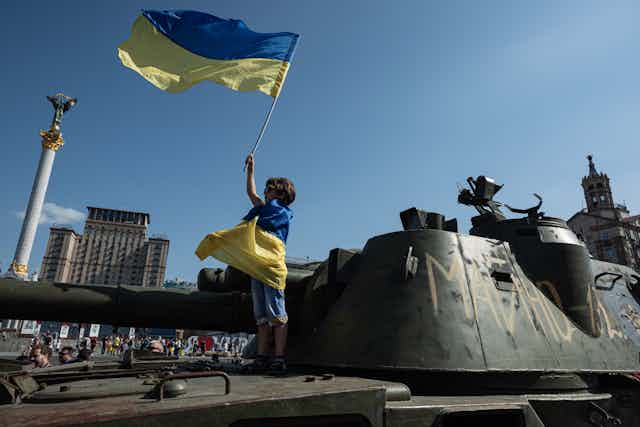 A small child is wrapped in a blue and yellow flag, and holds an identical flag in the air. He stands on top of a military vehicle. 