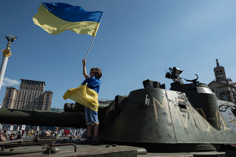 A small child is wrapped in a blue and yellow flag, and holds an identical flag in the air. He stands on top of a military vehicle.