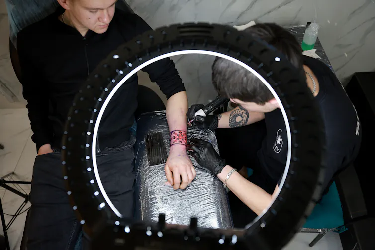 A man in a black shirt holds his arm on a black table covered in plastic, while another person appears to tattoo his arm with a tattoo gun.