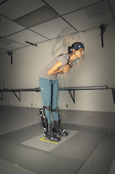 a standing young woman wearing mechanical devices on her lower legs bends forward at the waist and robot boots