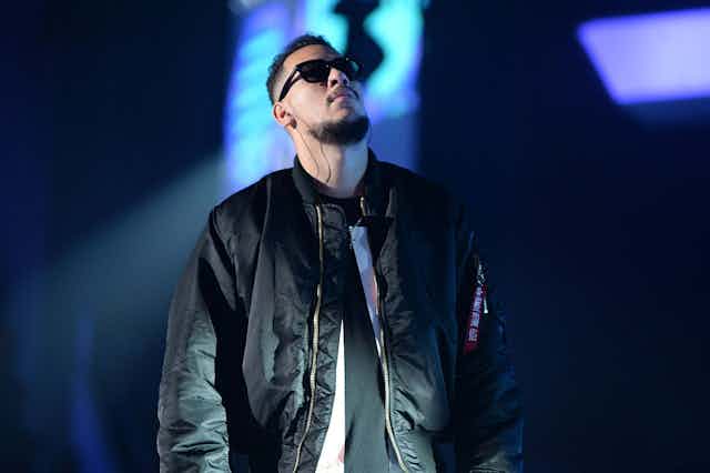 A man in a black leather jacket and in shades stands on a stage, a screen behind him. He looks up to the heavens.