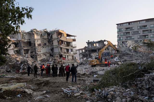 a group of men stand among the rubble of buildings in various stages of collapse