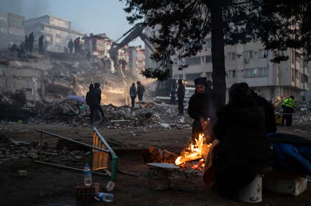 People huddle around a fire as rescuers search a destroyed building behind them in Kahramanmaras, southeastern Turkey.