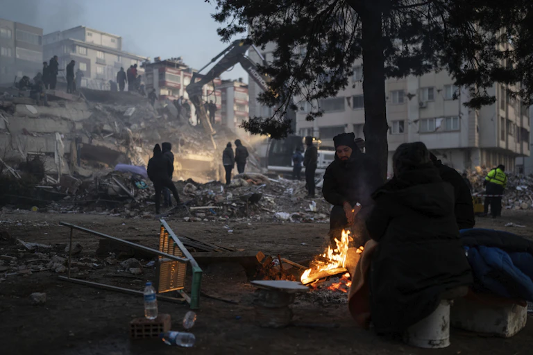 People huddle around a fire as rescuers search a destroyed building behind them in Kahramanmaras, southeastern Turkey.