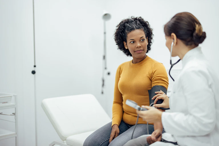 A doctor wearing a stethoscope and white coat measures the blood pressure of an African American woman in a medical exam room.