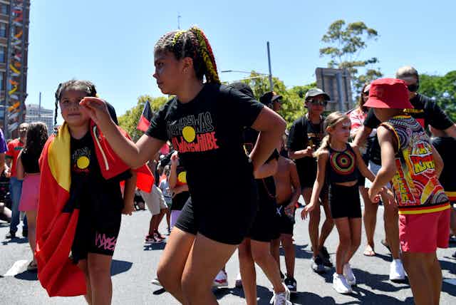 A young First-Nations girl, wearing a t-shirt displaying a design of the Aboriginal flag with text reading "Always was, always will be", dancing in a crowd of other young people on a sunny day 