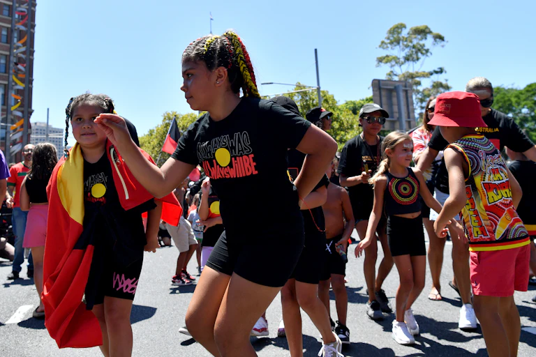 A young First-Nations girl, wearing a t-shirt displaying a design of the Aboriginal flag with text reading "Always was, always will be", dancing in a crowd of other young people on a sunny day