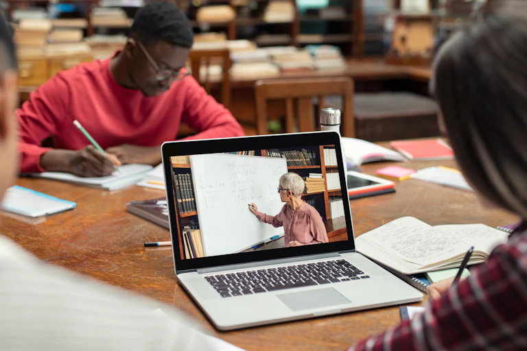 Two people working in a library; one, in a red shirt and wearing glasses, is making notes on a piece of paper. The other, their back to the camera, wears a plaid shirt and takes notes while watching an online lecture on their laptop