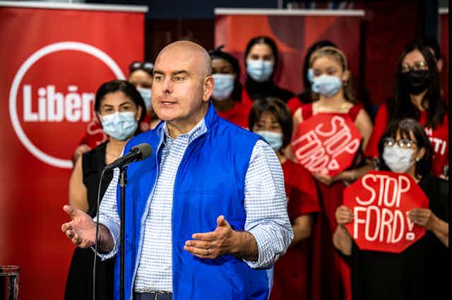 A bald man in a blue vest speaks in front of women in masks who carry signs that read Stop Ford.