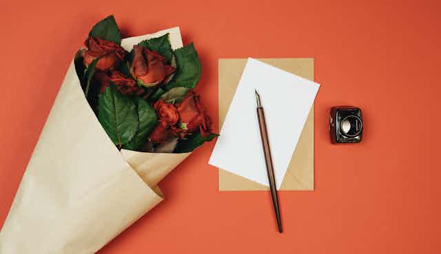 A bunch of red roses lies on a table next to a blank piece of paper, quill and ink-or.