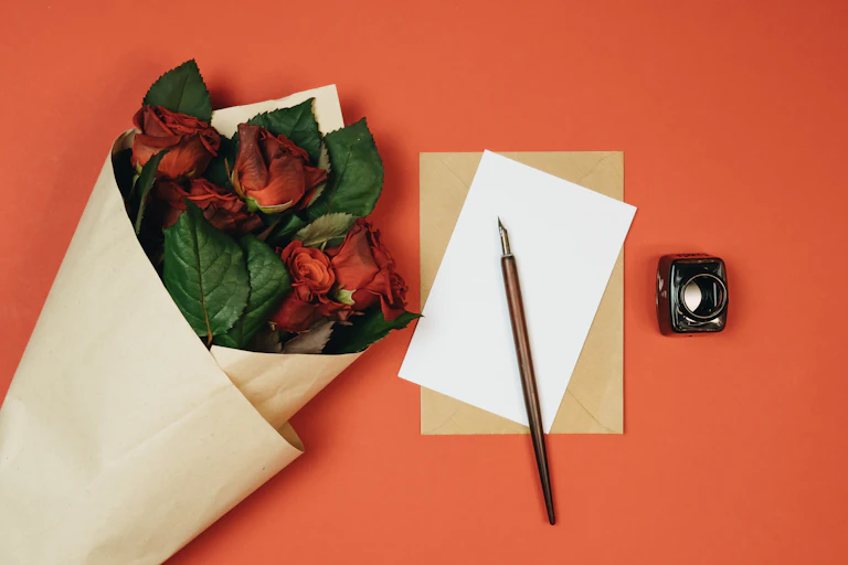 A bunch of red roses lies on a table next to a blank piece of paper, quill and ink-or.