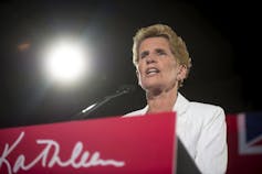 A woman in a white jacket with short dark blond hair speaks into a microphone.