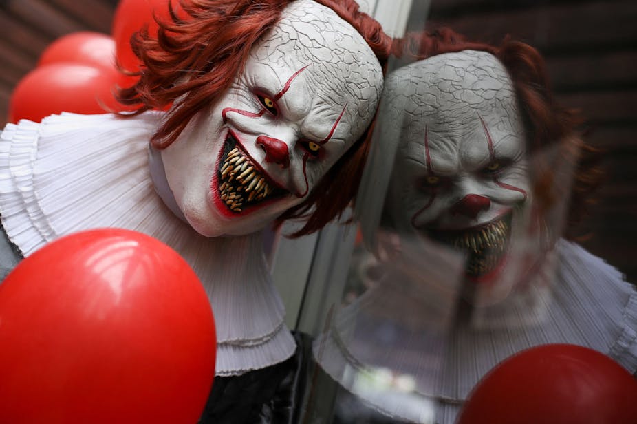 A sinister looking clown with red hair, white face, yellow eye and huge, sharp teeth leans against a window, creating a reflection.