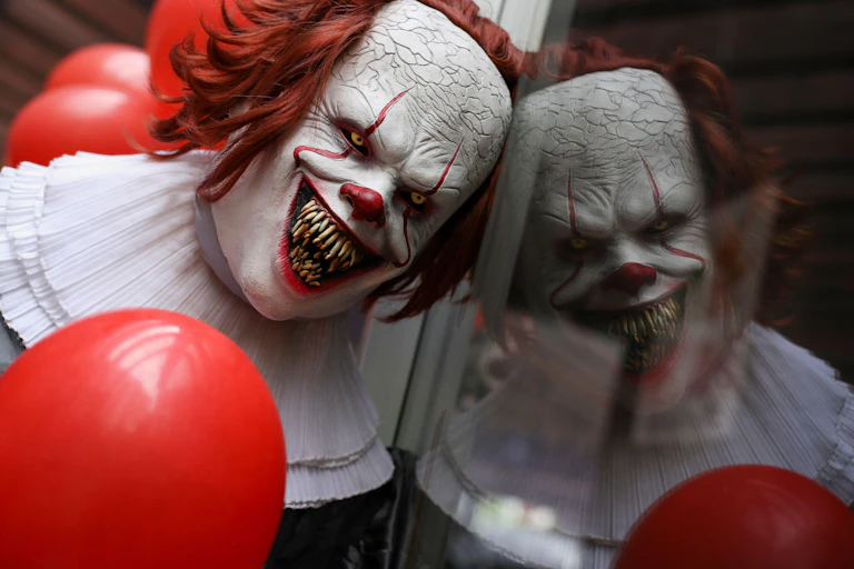 A sinister looking clown with red hair, white face, yellow eye and huge, sharp teeth leans against a window, creating a reflection.