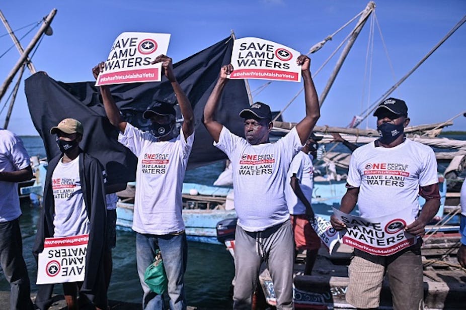 Four men holding up signs written 'Save Lamu Waters' in front of an ocean with boats in the background