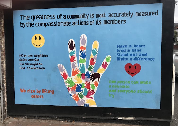 A large, handmade sign with a blue background and a colourful picture of a hand surrounded by quotes about community, neighbours and supporting each other.