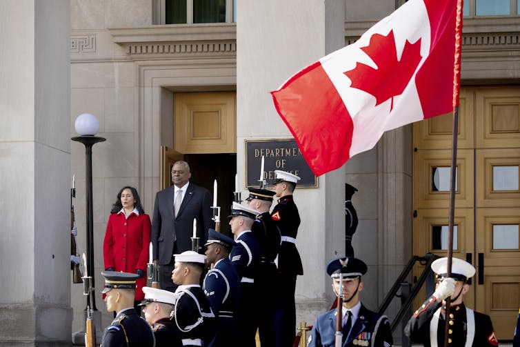 A Black man in a suit stands next to a dark-haired woman in a red coat, a Canadian flag in the foreground and a military band next to them.