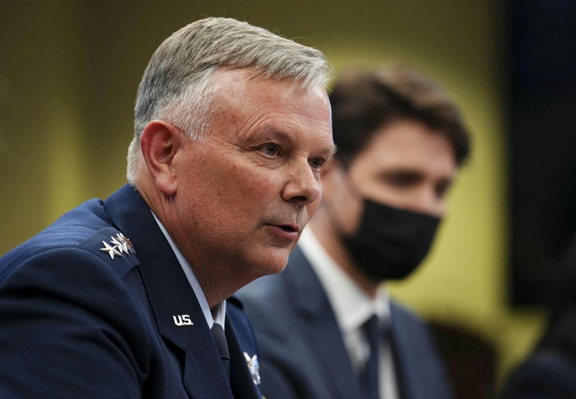 A grey-haired man in a military uniform speaks while a dark-haired man is seen behind him wearing a face mask.