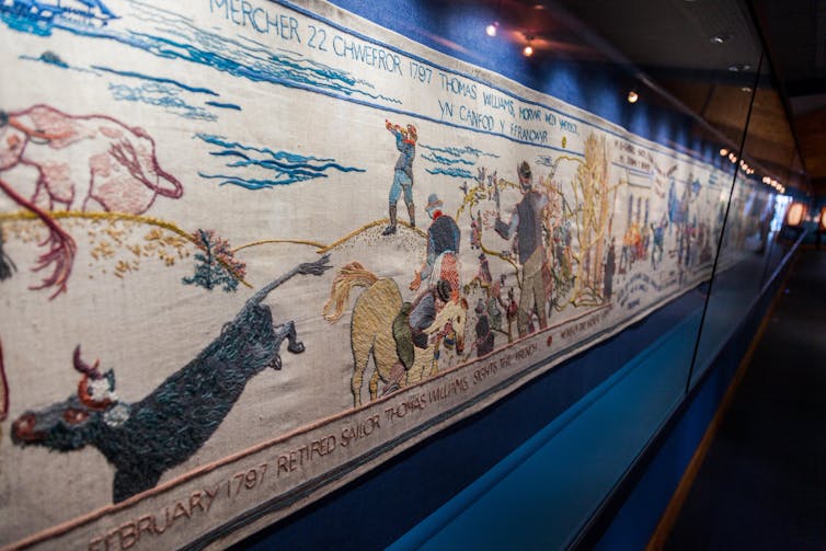 A very long tapestry with fine needlework displayed behind glass.