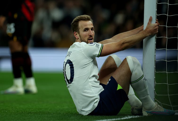 Harry Kane sits on the pitch holding onto a goalpost, looking directly into the camera.