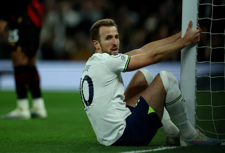 Harry Kane sits on the pitch holding onto a goalpost, looking directly into the camera.