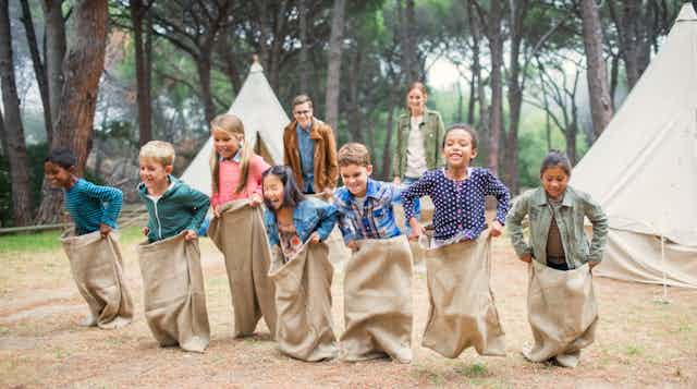 Seven smiling kids participate in a sack race as two adults look on.