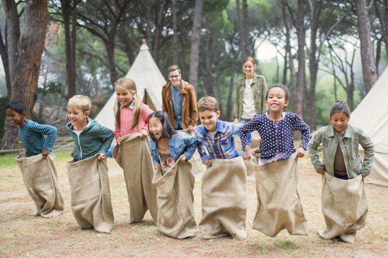 Seven smiling kids participate in a sack race as two adults look on.