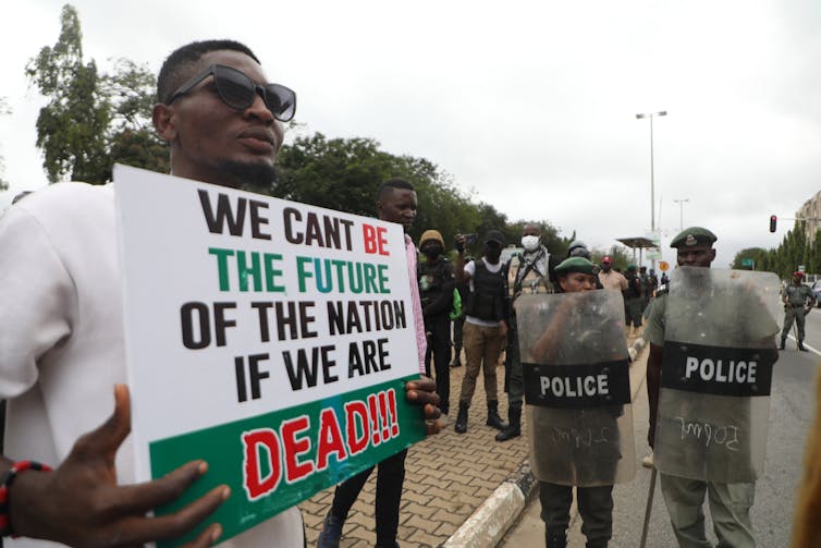 Man in sunglasses holds protest sign while police in riot shields walk nearby