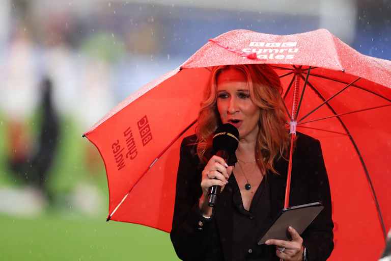 A woman in a black jacket holds a microphone in her right hand. In her left hand, she holds a red umbrella with the words "BBC Cymru Wales" on it.