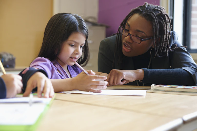 A teacher helps a student seated at a desk.