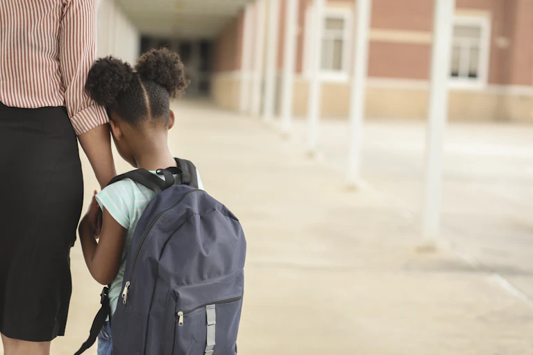 A girl with a backpack clings to her mother as her mother takes her to school.