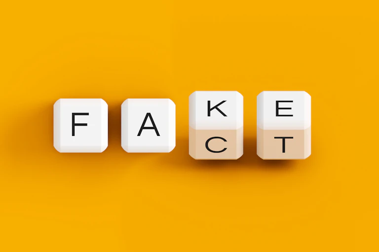Four cubes with letters on their sides spell out 'Fake' and 'Fact' against an orange backdrop.