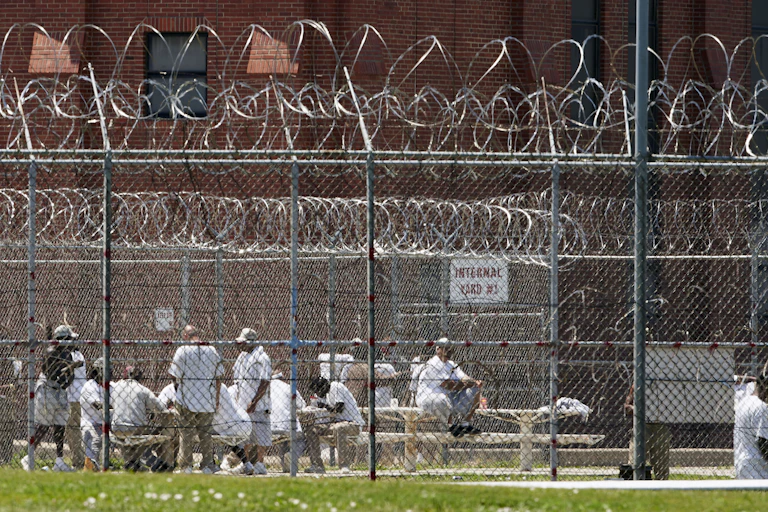 Several men wearing white t-shirts are seen behind a high wall covered with barbed wire.